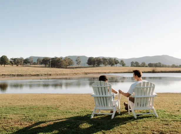 Couple seated in white Adirondack chairs beside the lake at The Lane Retreat, with mountains in the distance.